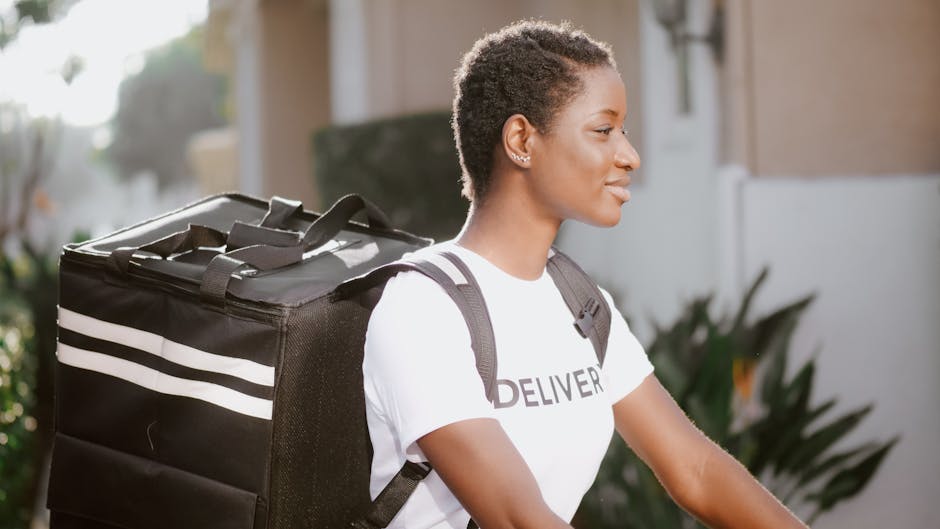 A smiling woman with short, curly hair is shown outdoors, standing next to a building with greenery in the background. She is wearing a white t-shirt with the word 'DELIVERY' printed on it and is carr