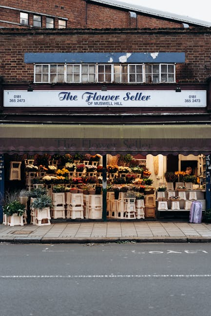 The storefront of The Flower Seller of Muswell Hill features a variety of colorful floral arrangements displayed on white stands and shelves inside the shop, with fresh flowers including yellow, pink,