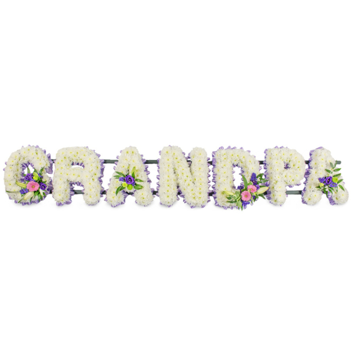 Displayed against a simple, pure white background, a bespoke floral arrangement forms the word "GRANDPA" in bold, sculpted letters designed for a funeral or memorial service. Each individual letter is tightly packed with clusters of crisp white chrysanthemums or similar small, multi-petalled flowers, creating a luxurious, cushion-like texture that suggests comfort and gentle remembrance. A delicate edging of tiny lilac and soft purple blossoms traces the outline of every letter, adding a subtle halo of colour that crisply defines the name while remaining soft and understated. The letters are aligned along a slim, dark green horizontal frame, which peeks through discreetly and hints at the sturdy structure that allows the tribute to stand proudly. Within the white petals of selected letters, small yet vivid accent sprays add depth and meaning: on the 'G' and first 'A' to the left, you can see slender white lily-like flowers combined with bright pink and fuchsia blooms, hints of deeper purple, and fresh green foliage; the 'D' and one of the later 'A's echo this combination, occasionally featuring lime-green button chrysanthemums that bring a lively, modern twist. The overall palette remains predominantly white and lilac, with these bolder touches drawing the eye and suggesting cherished memories or favourite colours of the grandparent being honoured. The scene feels calm and dignified, reminiscent of a respectful farewell in an Old Malden chapel or at a graveside near local green spaces. Soft, even lighting reveals every petal and contour, underscoring the care, skill, and affection that go into creating such a personalised name tribute for a much-loved grandfather.